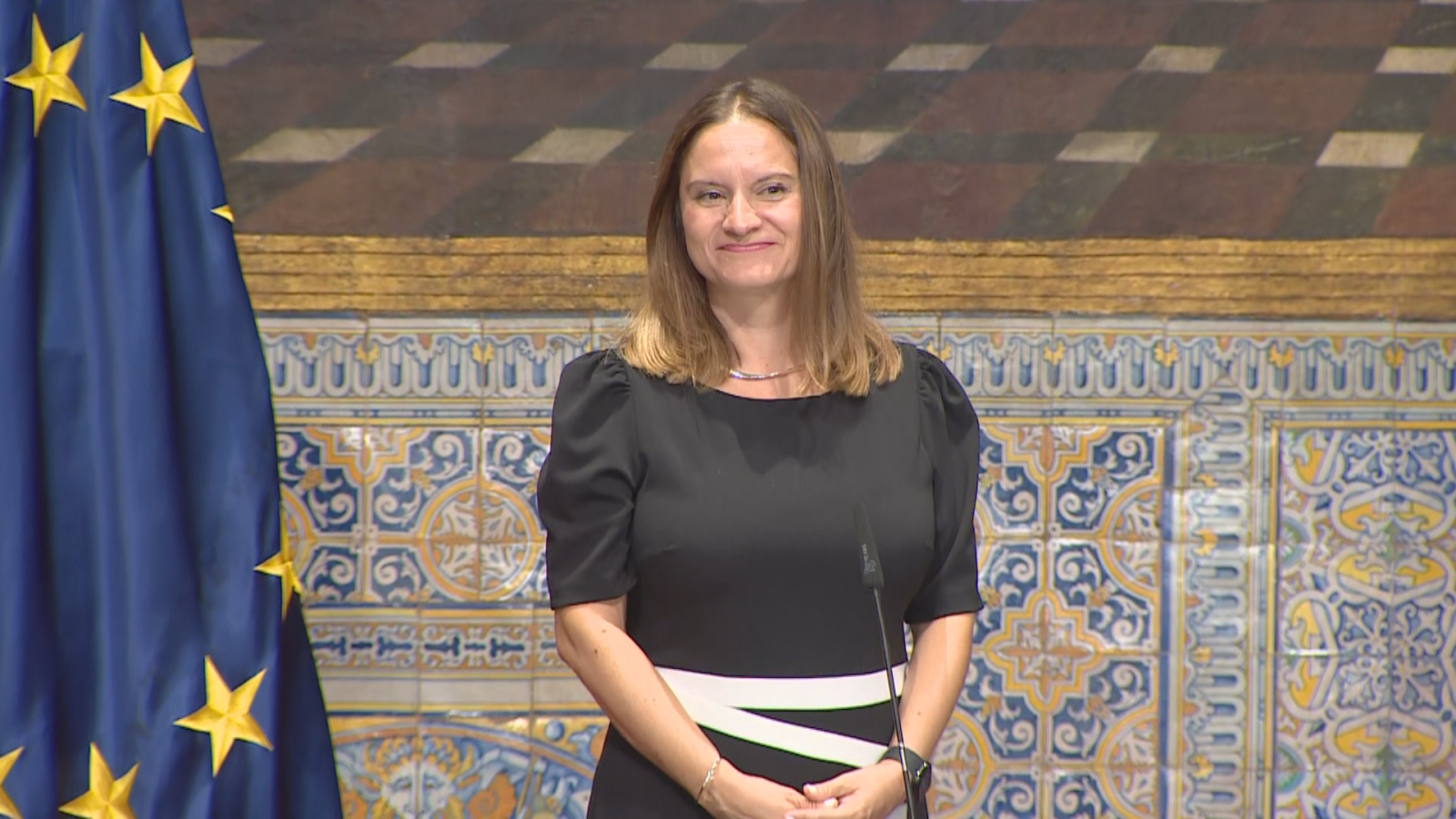 Aitana Ferrer, presentadora de "L'informatiu - Comunitat Valenciana", posa con un vestido negro y blanco, ante azulejos valencianos y la bandera de la UE.