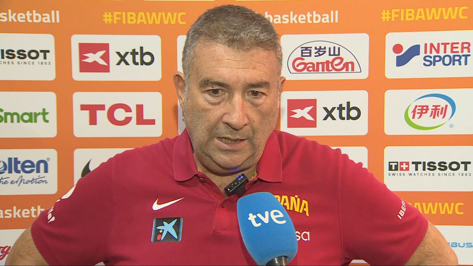 Miguel Méndez, seleccionador de baloncesto femenino español, en primer plano con polo rojo, logos de Nike y CaixaBank, y "ESPAÑA" en amarillo. Micrófono de TVE y fondo de patrocinadores FIBA.