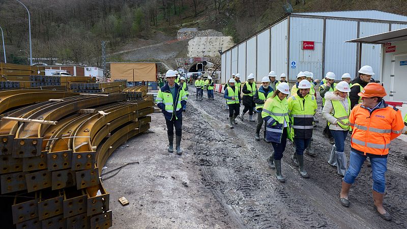 Grupo de personas con equipo de seguridad en obra, inspeccionando vigas metálicas y la entrada de un túnel. Se observa el logo de "Acciona" en un edificio.