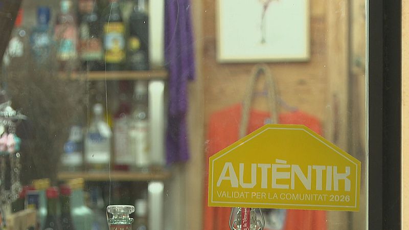 Escaparate de comercio con pegatina "AUTÈNTIK VALIDAT PER LA COMUNITAT 2026". Interior con botellas, textiles y cuadro.