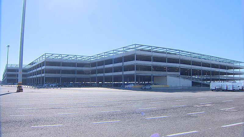 Gran silo vertical de automóviles en construcción en el Puerto de Santander, con vehículos estacionados y furgonetas blancas en la explanada.