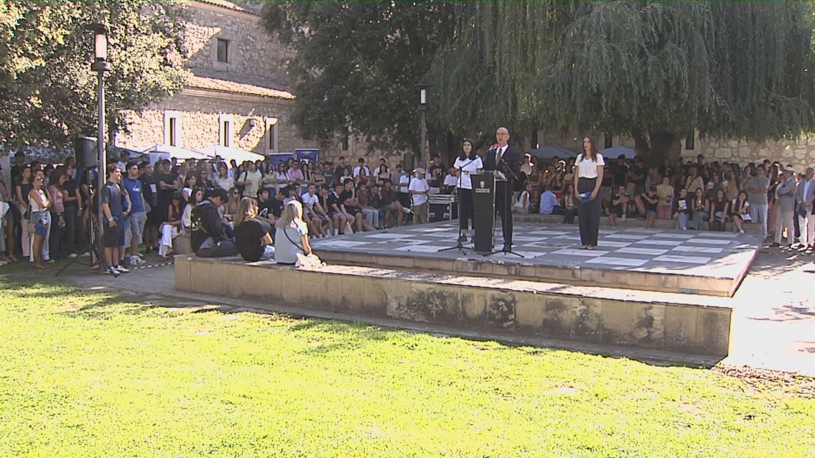 Escenario al aire libre con oradores dando la bienvenida a estudiantes universitarios. Fondo de campus con edificios de piedra y árboles. Audiencia joven congregada.