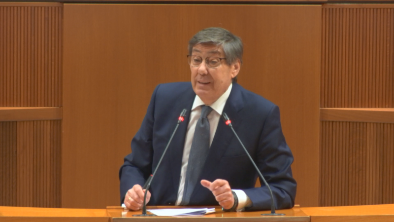 Arturo Aliaga, político español, habla en un evento formal con dos micrófonos frente a él. Viste traje oscuro y gesticula con las manos.