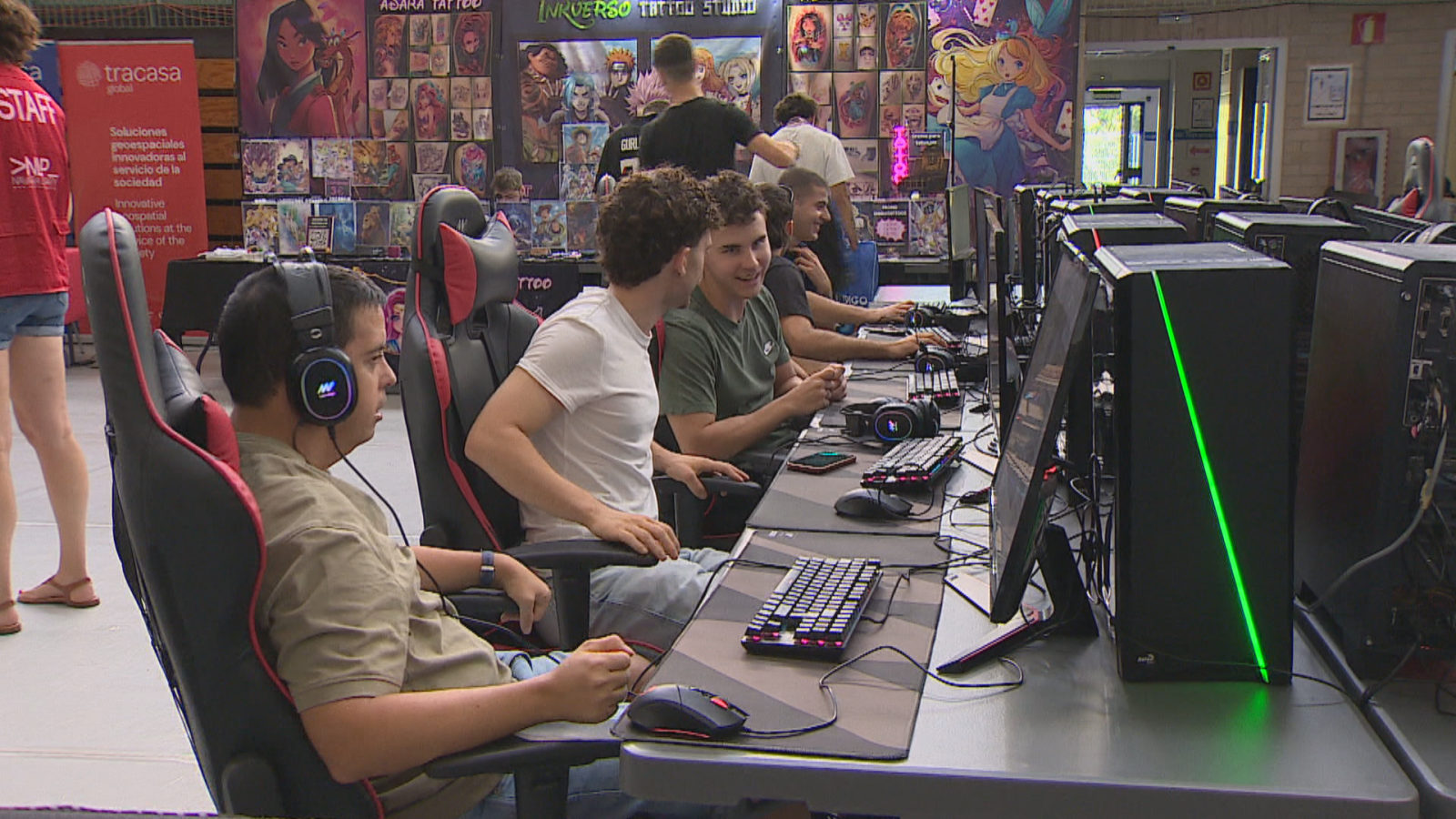 Jóvenes en una LAN Party, rodeados de equipos informáticos con luces LED verdes, auriculares y banners de "tracasa" e "INVERSO TATTOO STUDIO".