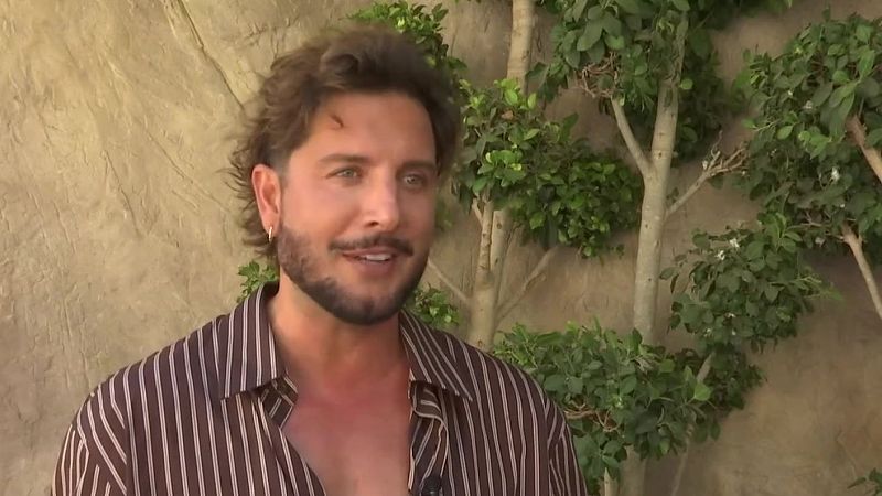 Omar Montes, cantante español, en primer plano con cabello rizado, barba y camisa de rayas marrones y beige. Fondo de pared texturizada y vegetación con flores blancas.