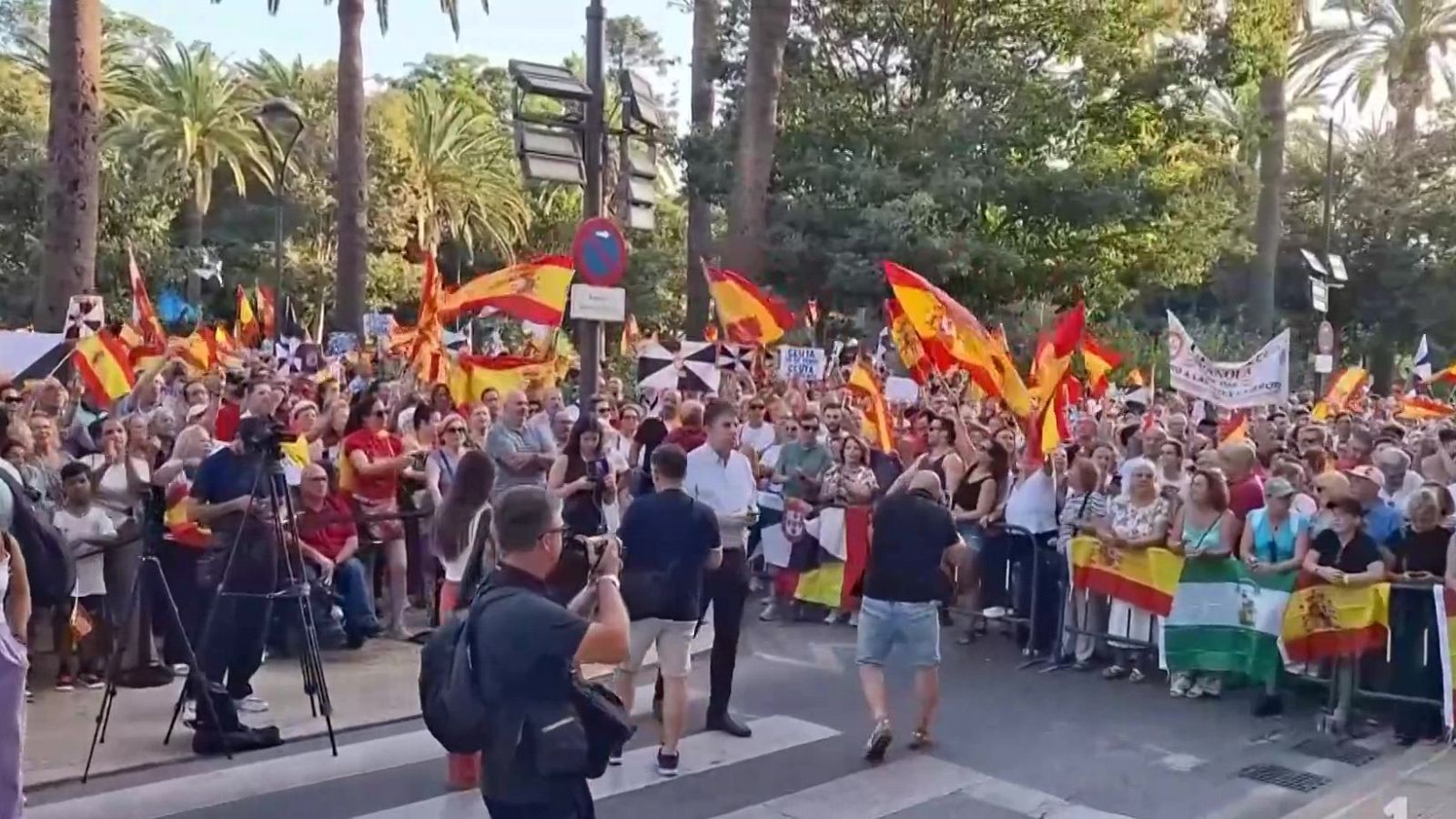 Manifestación política en Andalucía con Juanma Moreno Bonilla, rodeado de banderas de España, Andalucía y símbolos históricos. Personal de medios cubre el evento.