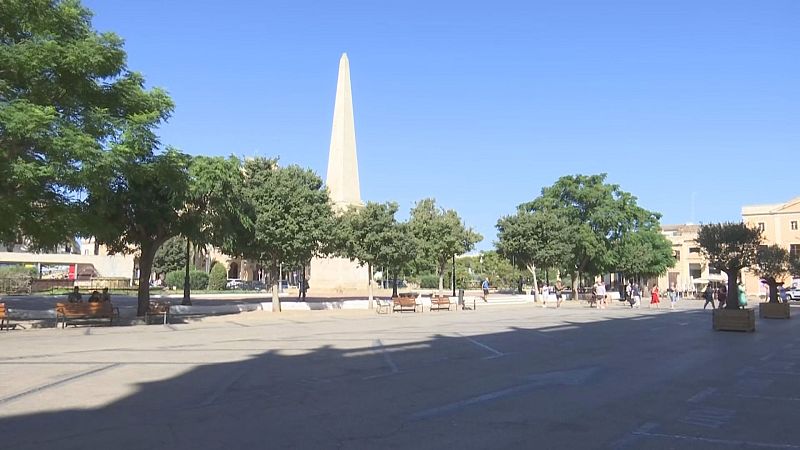 Vista de la Plaza del Borne en Ciutadella de Menorca, con el Obelisco, árboles, edificios mediterráneos y personas transitando bajo un cielo soleado.