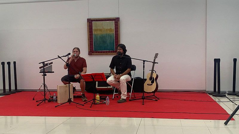 Dúo musical en escenario con alfombra roja: un percusionista/vocalista y un guitarrista, ante un fondo blanco con un cuadro abstracto.