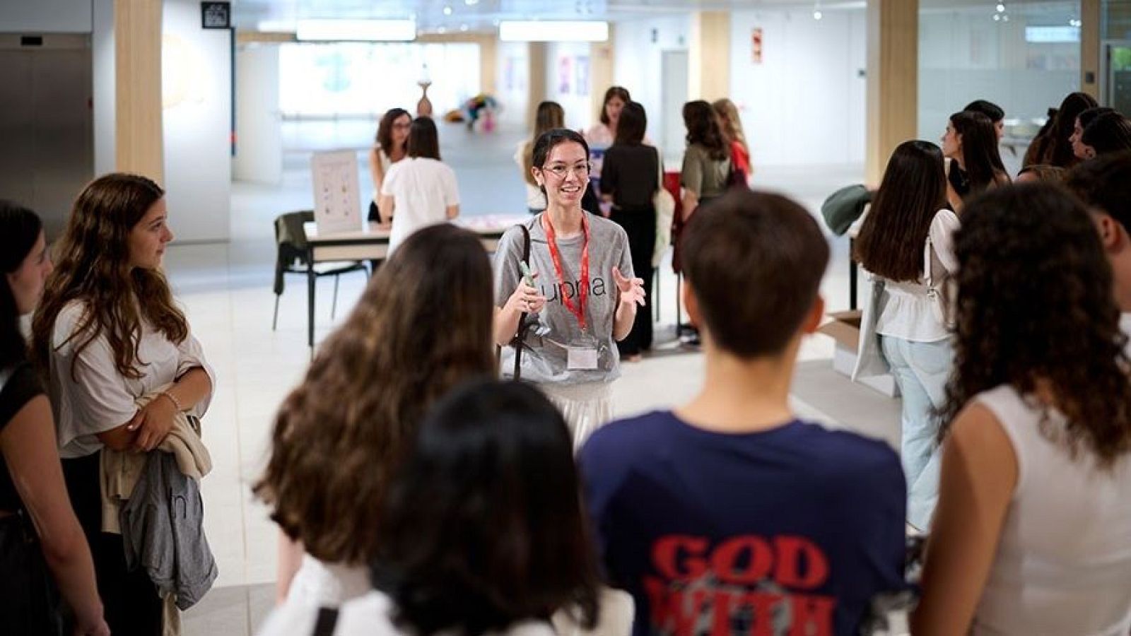 Jóvenes en un evento informativo en la UPNA. Una oradora con camiseta "upna" explica algo a un grupo de estudiantes en un edificio moderno y luminoso.