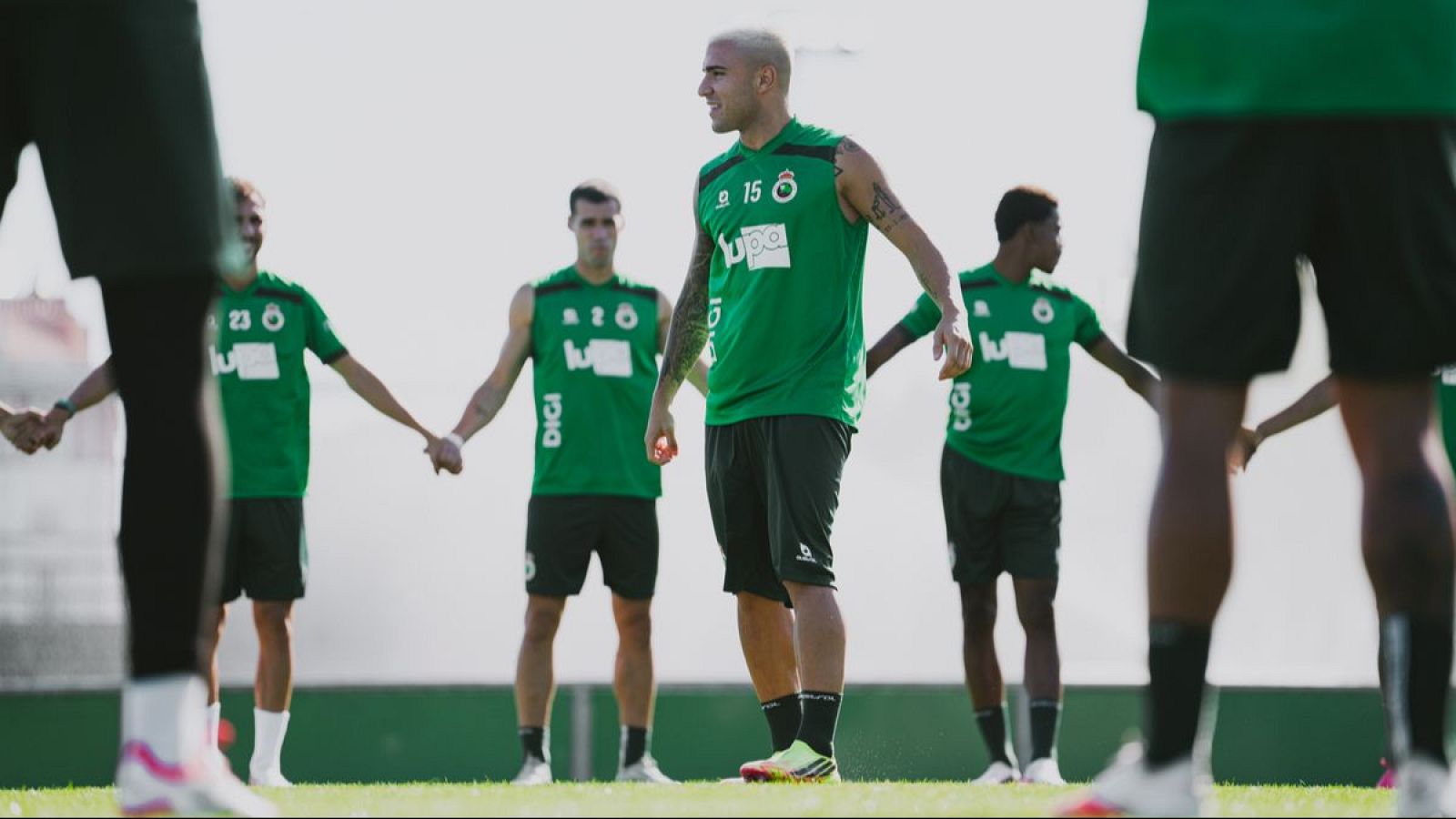 Jugadores del Real Racing Club de Santander entrenan en un día soleado, formando una cadena sobre el césped con la equipación del equipo.