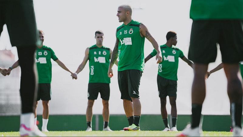 Jugadores del Real Racing Club de Santander entrenan en un día soleado, formando una cadena sobre el césped con la equipación del equipo.