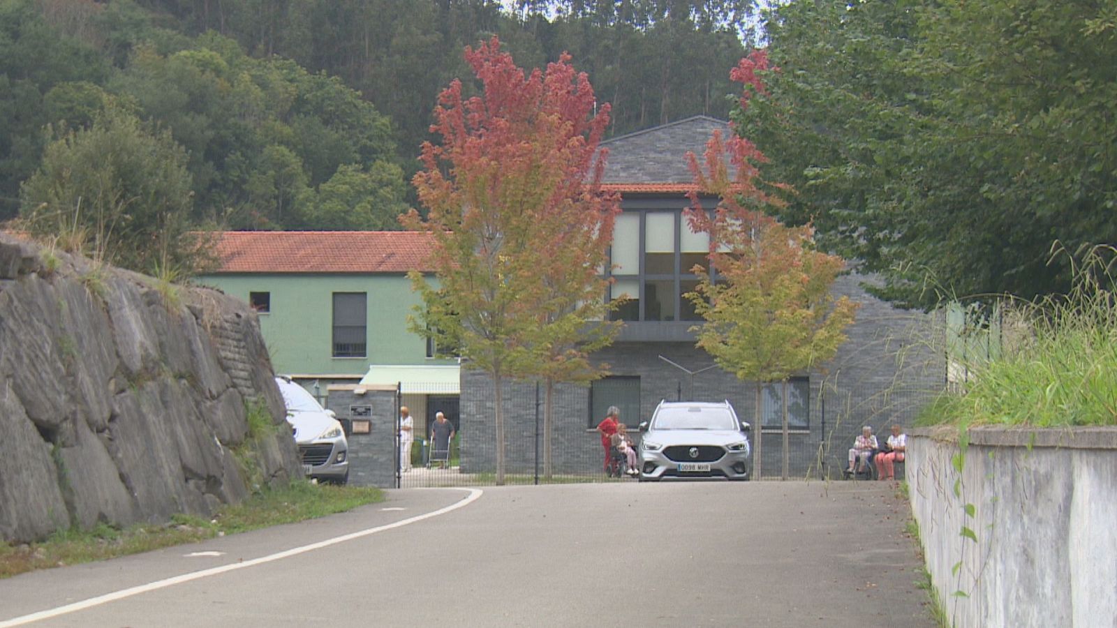 Vista exterior de la Residencia Valdáliga en Cantabria, con edificios modernos y clásicos, rodeada de naturaleza otoñal. Se observan vehículos y personas en el área de acceso.