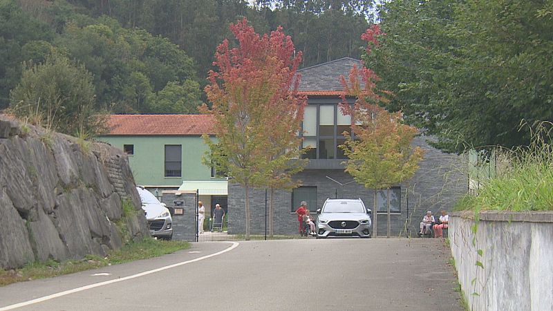 Vista exterior de la Residencia Valdáliga en Cantabria, con edificios modernos y clásicos, rodeada de naturaleza otoñal. Se observan vehículos y personas en el área de acceso.