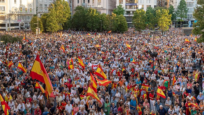 Vista aérea de una multitud masiva ondeando banderas de España en un espacio público urbano, con edificios y árboles al fondo.