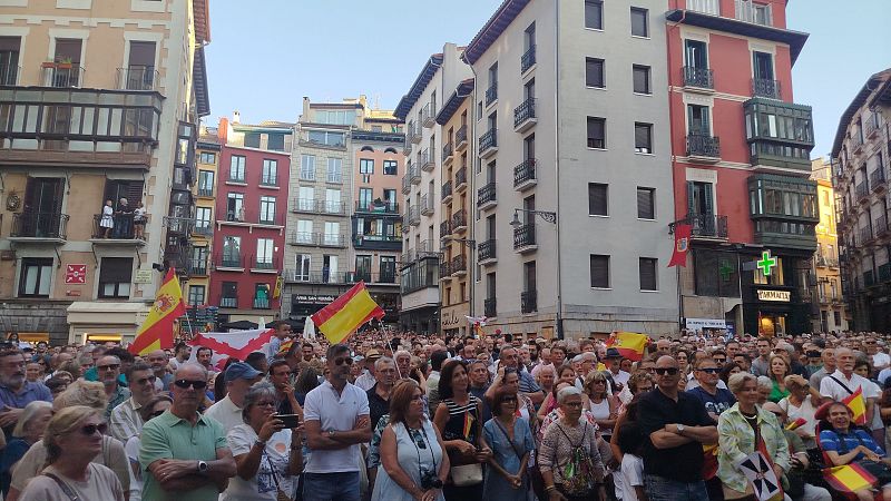 Multitud en plaza española al atardecer, ondeando banderas de España. Edificios tradicionales y farmacia al fondo. Luz dorada ilumina la escena.