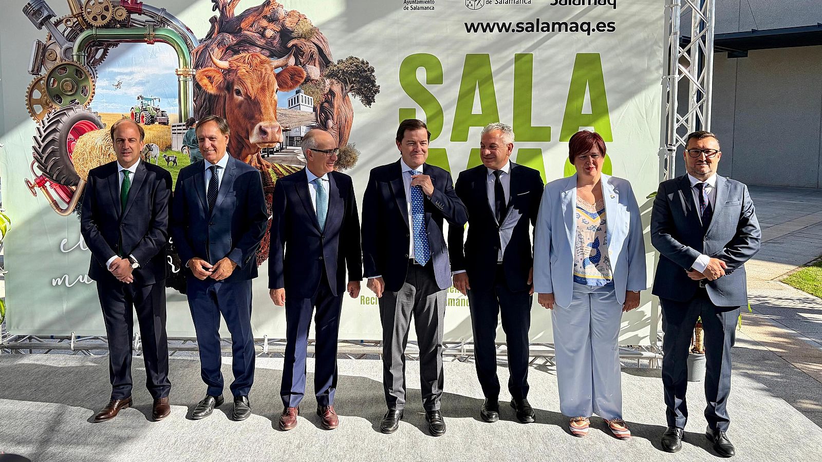 Grupo de siete personas, incluyendo a Alfonso Fernández Mañueco, posan frente a un panel de Salamaq con temática agrícola y ganadera.