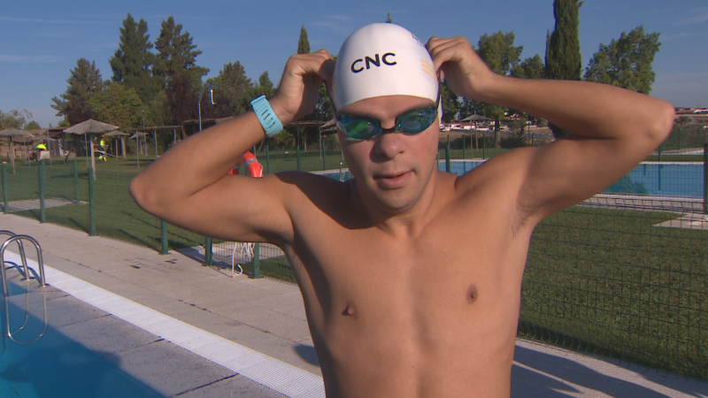 Hombre con gorro "CNC" y gafas azules se ajusta el gorro en el borde de una piscina, con reloj deportivo azul. Entorno soleado con césped y árboles.