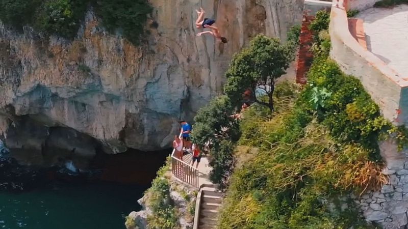 Joven en salto al mar desde acantilado con pasarela de madera y vegetación. Vista aérea en día soleado.
