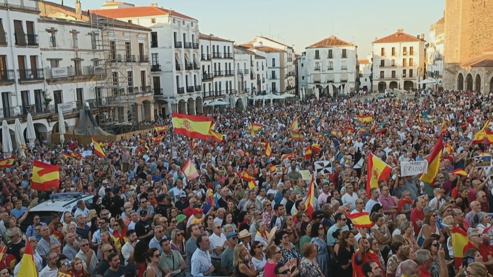 Vista aérea de miles de personas en la Plaza Mayor de Cáceres, España, con banderas españolas y pancartas. Se observa un vehículo policial y edificios históricos.