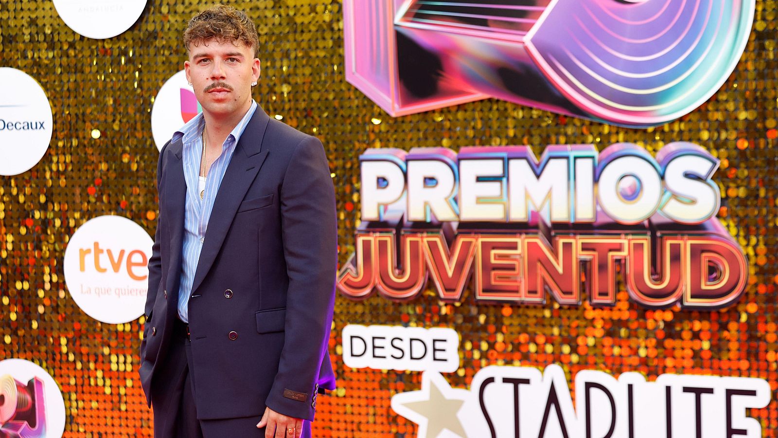 Quevedo, con traje azul y camisa de rayas, posa ante un fondo de lentejuelas doradas y ámbar con el logo "PREMIOS JUVENTUD" y patrocinadores como RTVE.
