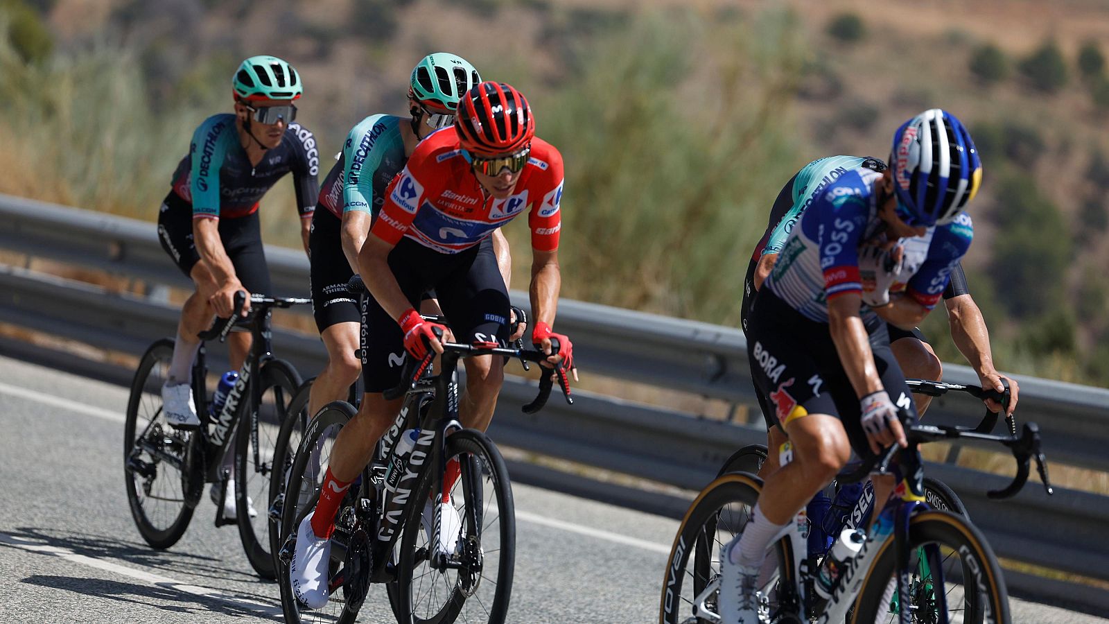 Ciclistas en plena competición, con maillots de "DECATHLON", "CARREFOUR" y "BORA", pedalean en una carretera soleada con fondo montañoso. Se aprecian detalles de cascos, gafas y bicicletas de marcas como "VANRYSEL" y "CANYON".