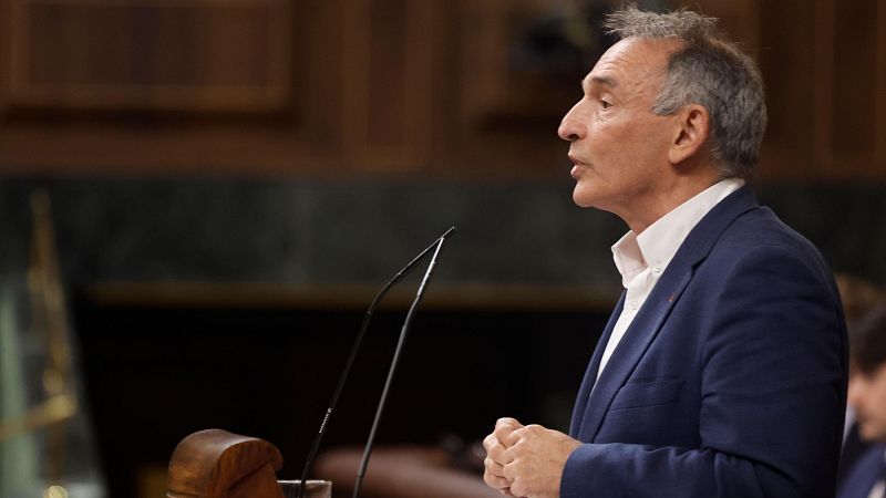 Primer plano de Patxi López, político español, en un discurso. De perfil, con traje azul y camisa blanca, habla con las manos entrelazadas. Fondo formal desenfocado.