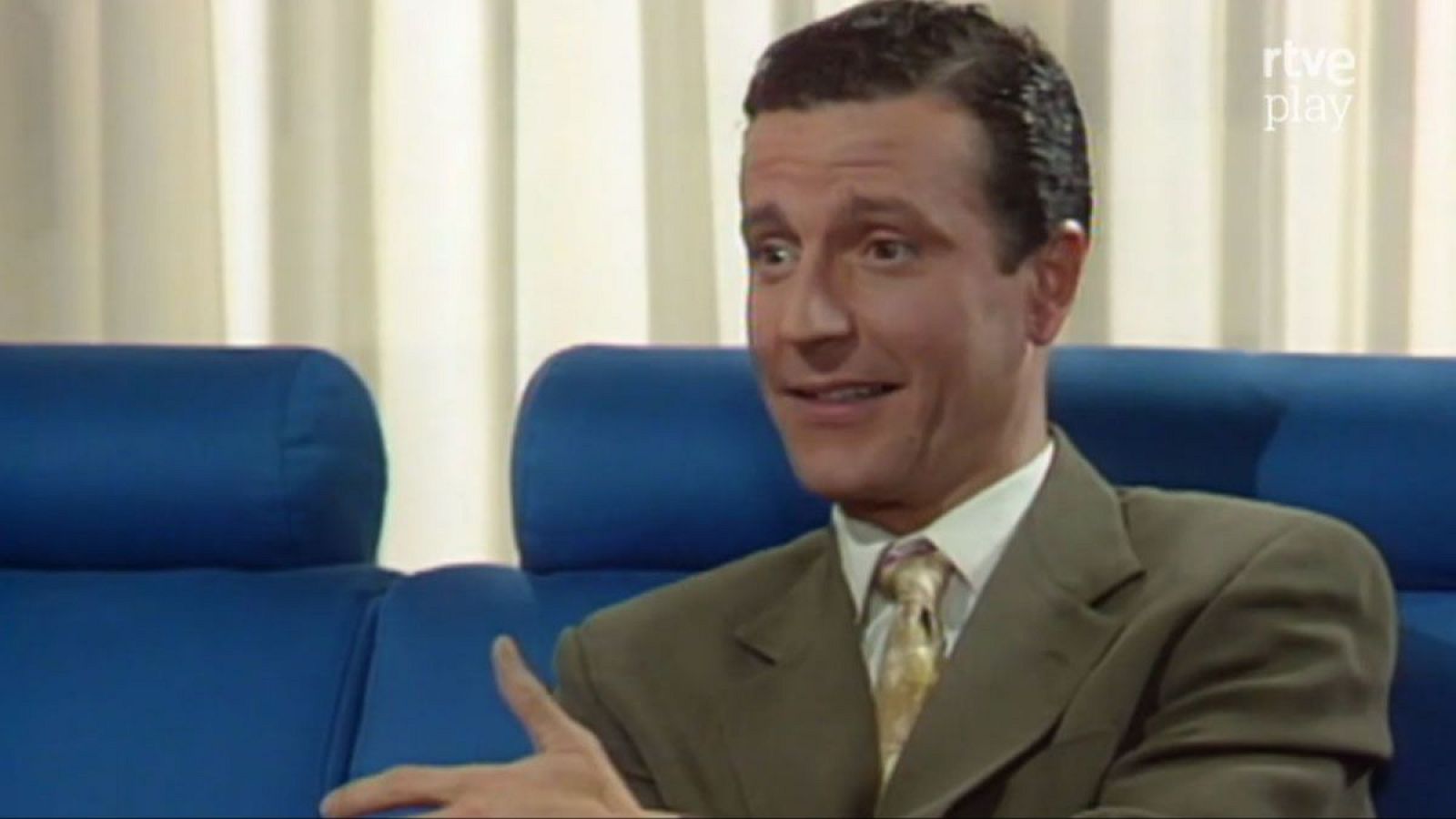 Alberto San Juan, actor español, en primer plano con traje verde oliva, camisa blanca y corbata dorada. Sonríe con la mano izquierda levantada. Fondo claro y sofá azul. Logo 'rtve play' visible.