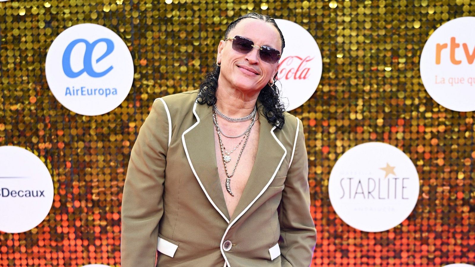 Elvis Crespo, con gafas oscuras y chaqueta verde oliva, sonríe ante un fondo de lentejuelas doradas y logotipos de patrocinadores.