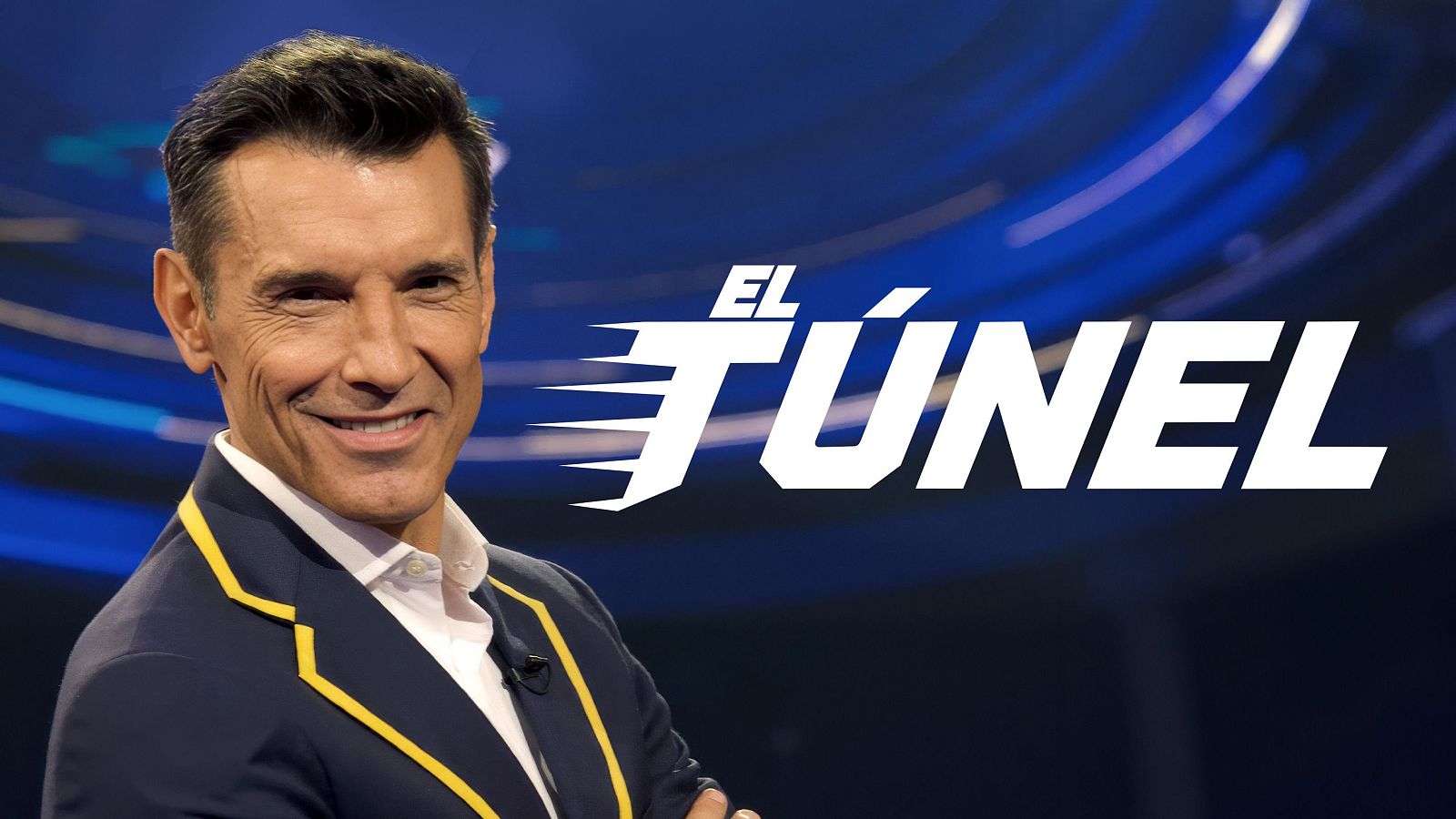 Jesús Vázquez sonríe a cámara con americana azul marino y ribete amarillo. Logo de "EL TÚNEL" a su derecha sobre fondo azul dinámico.