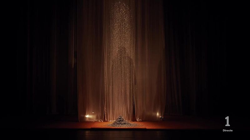 Escena teatral con cortinas beige, caída de partículas brillantes y un montículo cónico en el suelo. Indicativo de canal '1 Directe' en la esquina inferior derecha.
