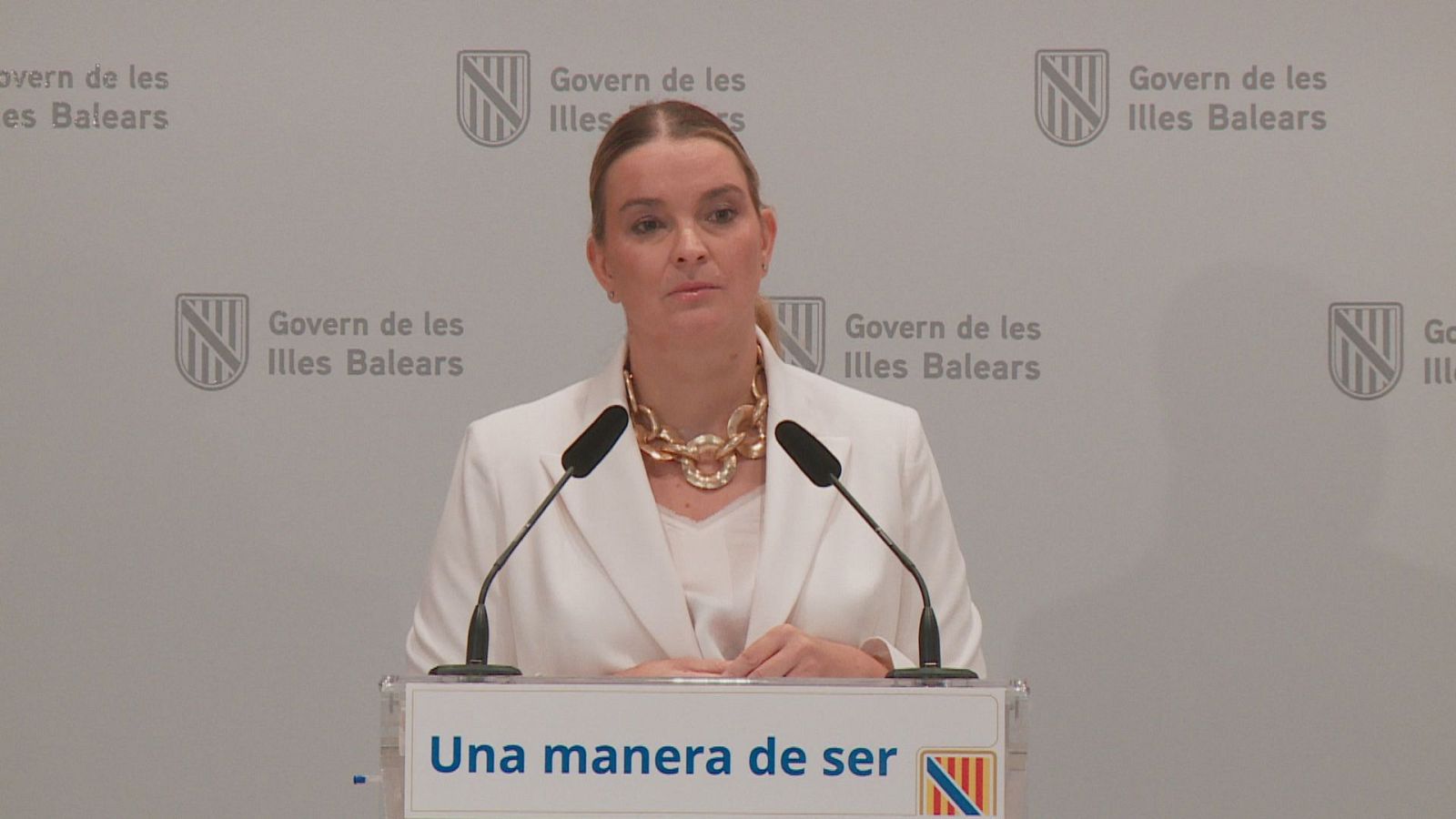 Marga Prohens, Presidenta del Govern de les Illes Balears, habla desde un atril con micrófonos. Lleva americana blanca, blusa de encaje y collar dorado. Fondo con logo del Govern.