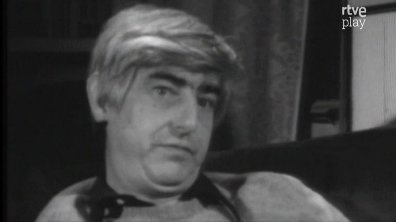 Primer plano en blanco y negro de Juan Benet, escritor español, con expresión seria. Se observa su cabello claro, cejas oscuras y vestimenta oscura. Fondo desenfocado y logo de RTVE Play.