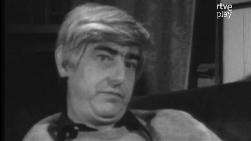 Primer plano en blanco y negro de Juan Benet, escritor español, con expresión seria. Se observa su cabello claro, cejas oscuras y vestimenta oscura. Fondo desenfocado y logo de RTVE Play.
