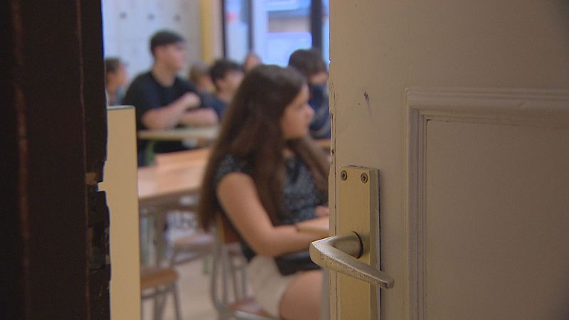 Joven de pelo castaño sentada en un pupitre en un aula, vista a través de una puerta entreabierta. Se aprecian otros estudiantes y mobiliario escolar.