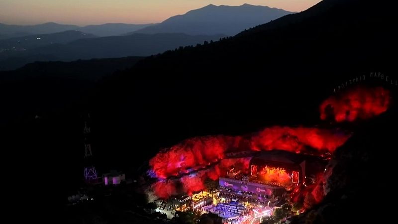 Festival nocturno en un anfiteatro montañoso con escenario iluminado, efectos rojos y público. Se lee "AUDITORIO PARQUE LL" en la ladera.