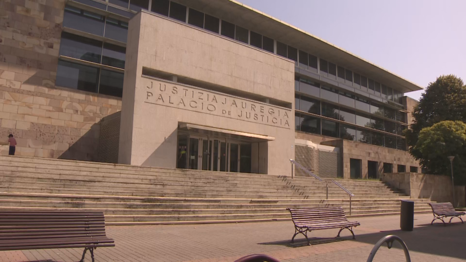 Fachada de un Palacio de Justicia con escalinata, inscripciones en euskera y español, bancos y vegetación bajo un cielo azul.