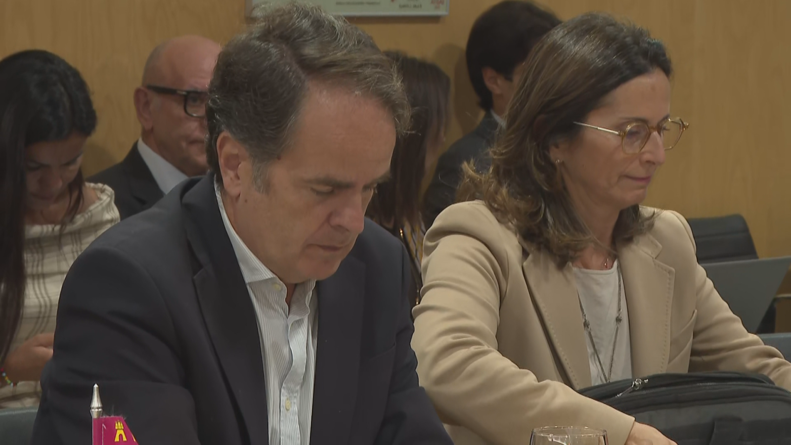 Fernando Gimeno Marín, con traje y camisa de rayas, se concentra en una reunión formal junto a una mujer con gafas. Se aprecian un estandarte rojo y un maletín negro en la escena.