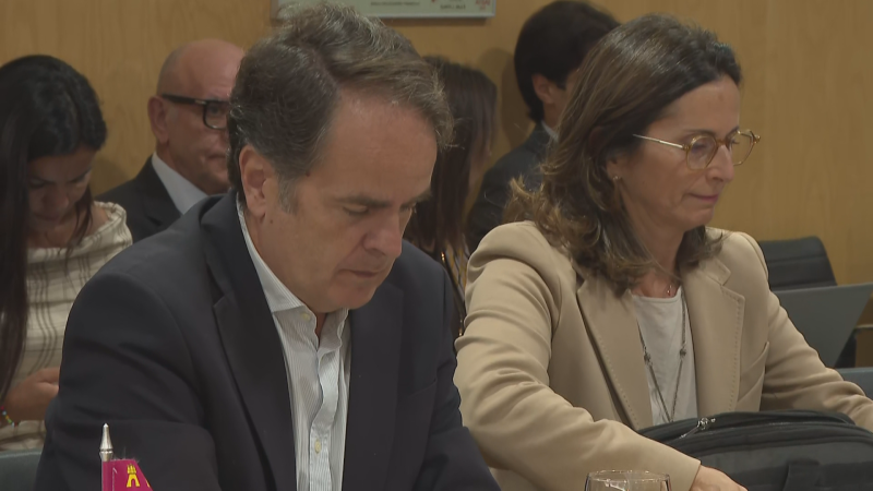 Fernando Gimeno Marín, con traje y camisa de rayas, se concentra en una reunión formal junto a una mujer con gafas. Se aprecian un estandarte rojo y un maletín negro en la escena.