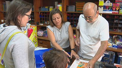 Persoal e clientes interact�an nunha tenda de material escolar, cun artigo laranxa en primeiro plano. Detr�s, estantes cheos de produtos e un expositor co texto "UCHES ANES".