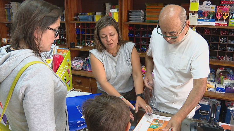 Persoal e clientes interactúan nunha tenda de material escolar, cun artigo laranxa en primeiro plano. Detrás, estantes cheos de produtos e un expositor co texto "UCHES ANES".