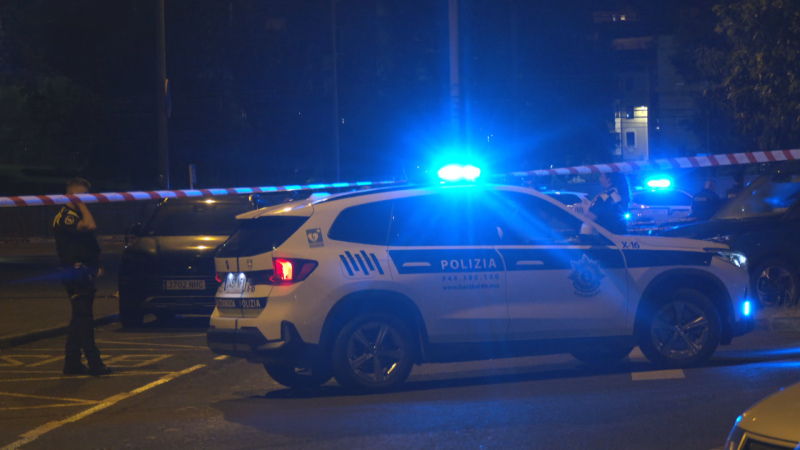 Vehículo policial "POLIZIA" con luces azules encendidas y cinta de seguridad roja y blanca en una calle urbana nocturna. Agente de policía presente.