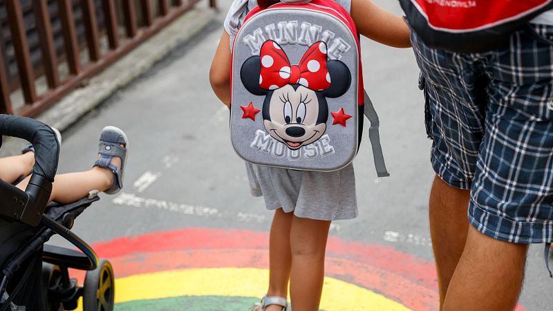 Mochila de Minnie Mouse con relieve, sandalias, pantalones a cuadros y un arcoíris en el suelo. Se aprecian las siglas 'RNE' en una mochila.