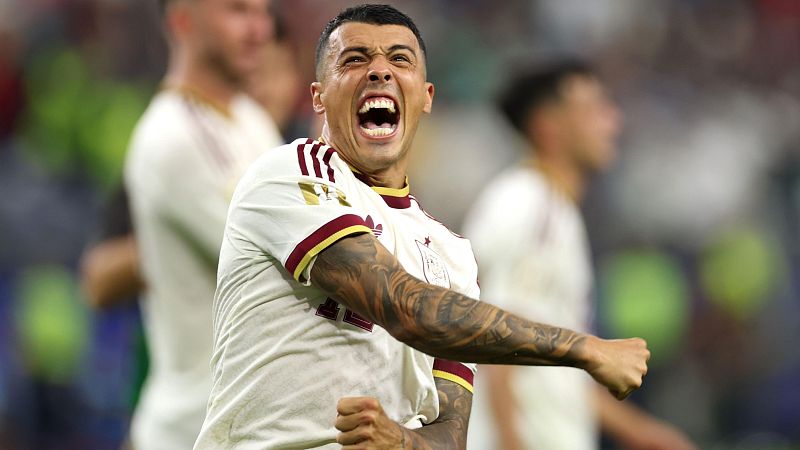 Pedro Porro celebra con euforia, boca abierta, ojos abiertos, camiseta deportiva blanca con detalles granates, dorados y Adidas, escudo con estrella. Fondo de estadio desenfocado.