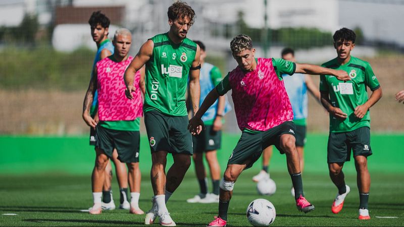 Jugadores de fútbol entrenan en un campo verde. Pol Moreno (camiseta verde, dorsal 5) y Germán Sánchez (camiseta rosa) participan en la sesión. Uno de ellos dribla el balón con destreza.