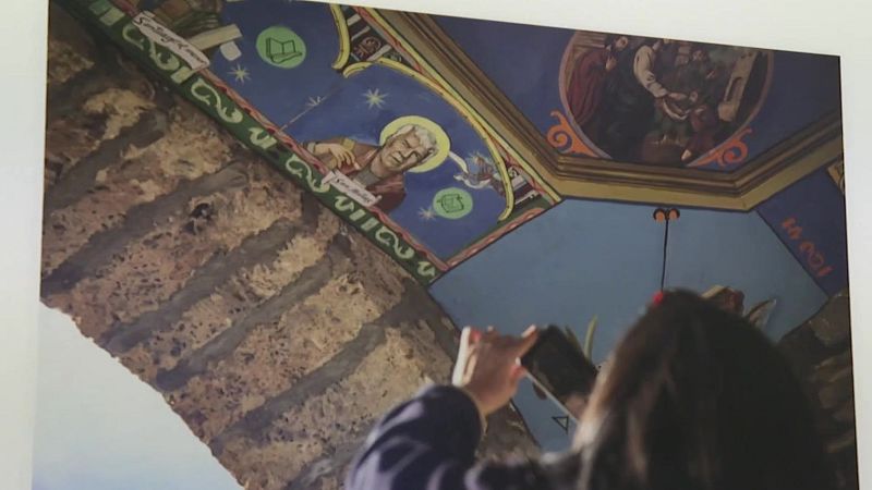 Persona fotografía obra de arte antigua en exposición. Destacan azules, dorados y figuras en la pintura, con posible referencia a Santiago.