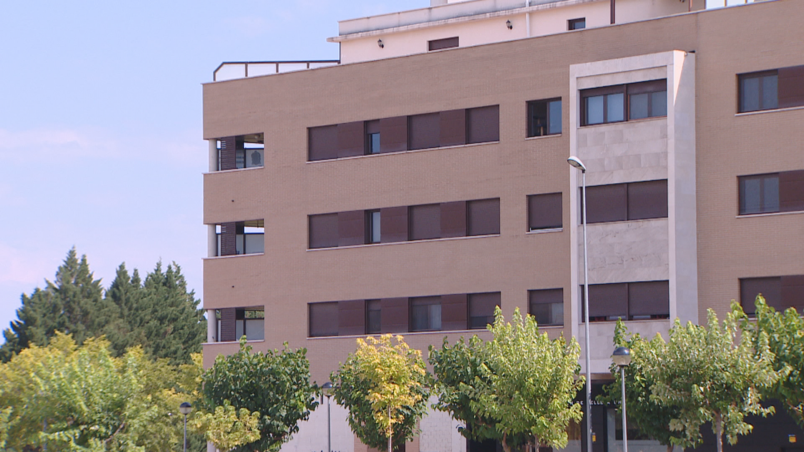 Edificio moderno beige y gris con ventanas oscuras y árboles verdes en primer plano, bajo un cielo azul despejado.