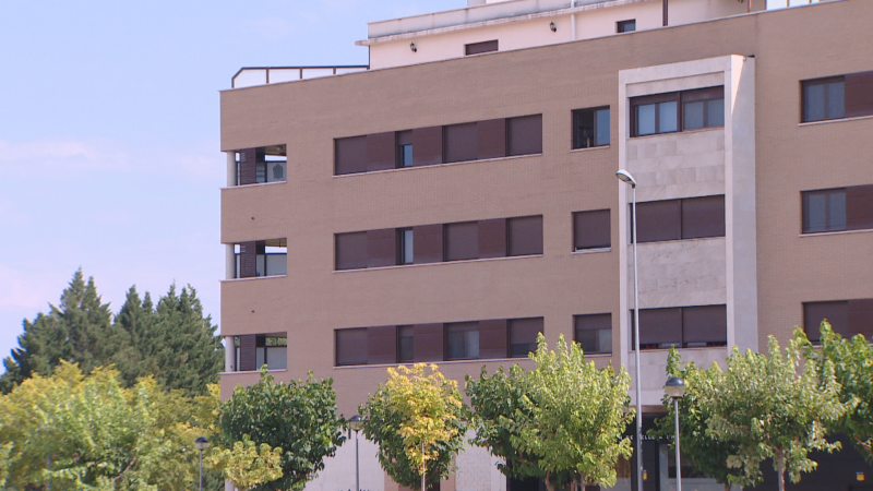 Edificio moderno beige y gris con ventanas oscuras y árboles verdes en primer plano, bajo un cielo azul despejado.