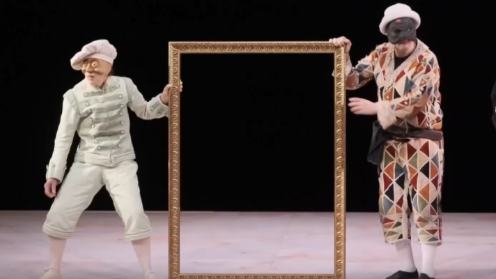 Arlecchino y un personaje enmascarado con traje claro presentan un gran marco dorado vacío en un escenario teatral de fondo negro, rememorando la Commedia dell'arte.