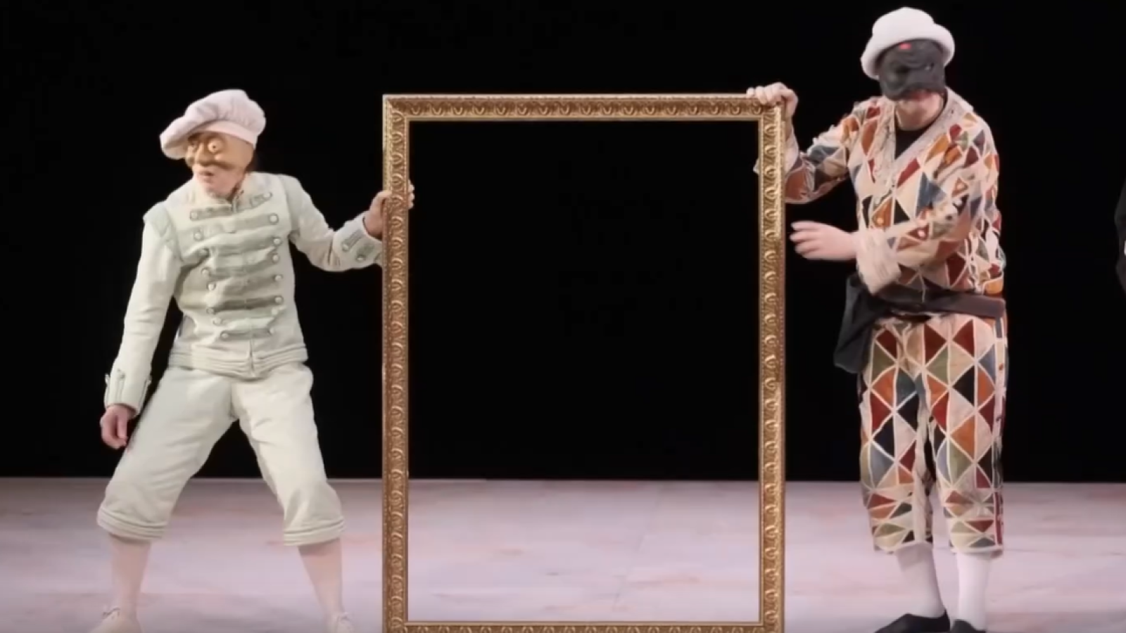 Arlecchino y un personaje enmascarado con traje claro presentan un gran marco dorado vacío en un escenario teatral de fondo negro, rememorando la Commedia dell'arte.