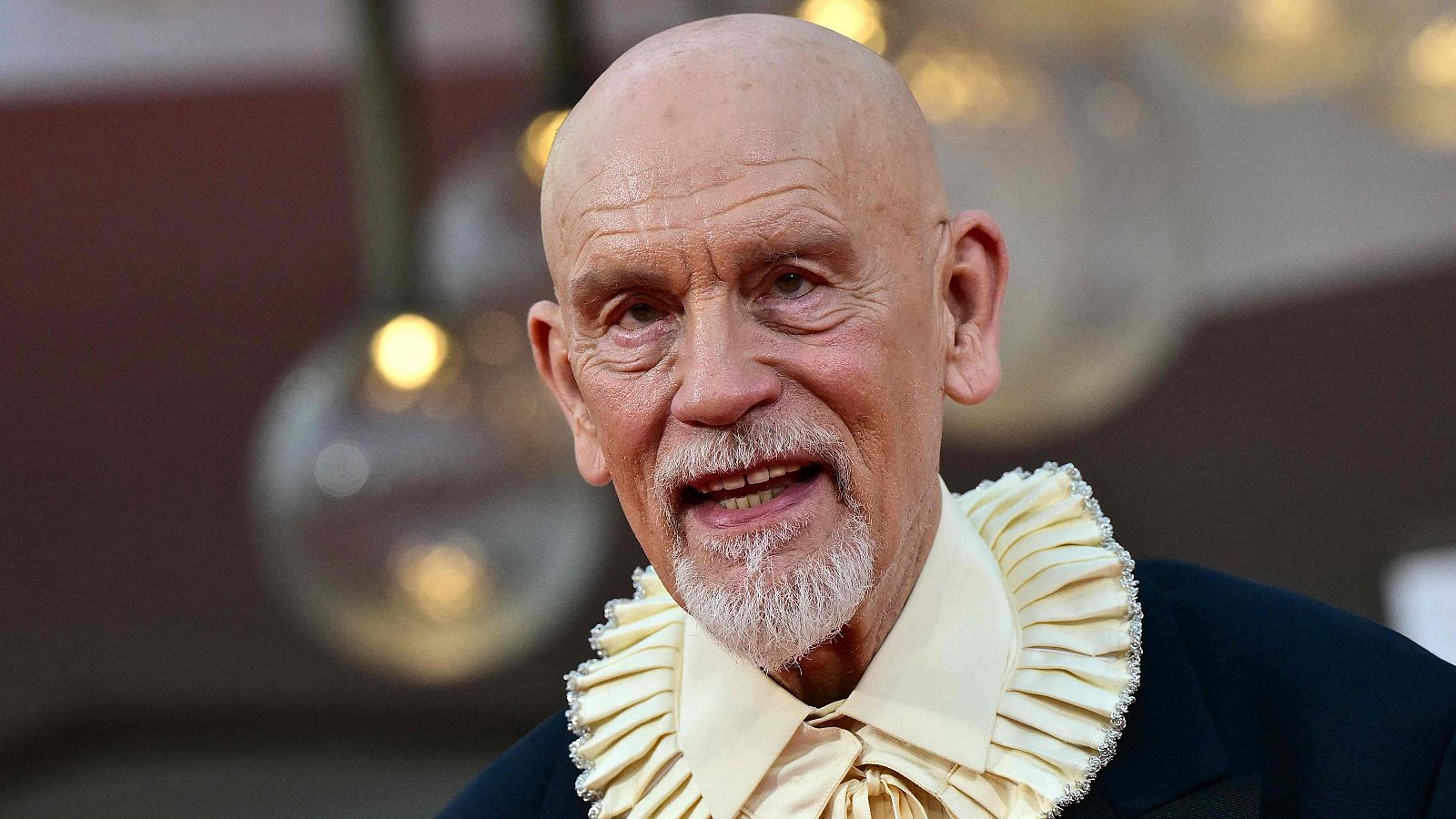 John Malkovich, hombre mayor con barba blanca y cuello de camisa voluminoso y adornado, sonríe levemente ante un fondo bokeh de luces cálidas.